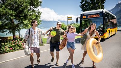 A group of young people with bathing utensils crossing a road by Lake Thun in front of a Postbus.