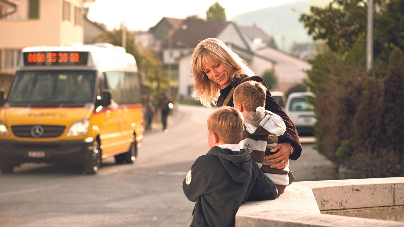 Eine Mutter wartet mit zwei Kindern an der Postauto-Haltestelle. Im Hintergrund ist ein Postauto zu sehen.