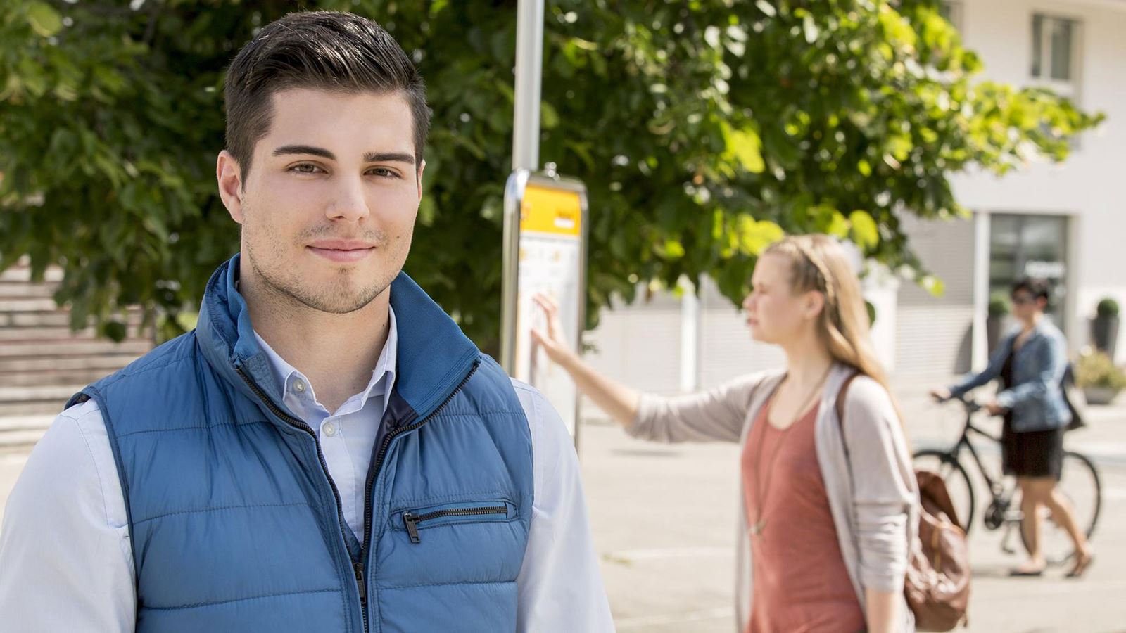 A man stands at a Postbus stop. Someone is looking at the timetable in the background.