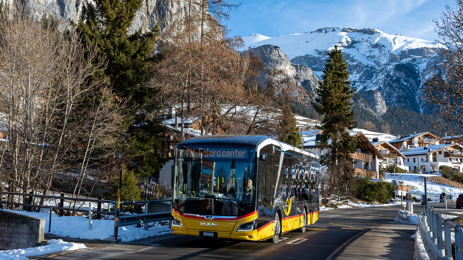 In a snowy Swiss Alpine landscape, the Postbus drives through a mountain village with traditional wooden houses and views of high snow-covered mountains in winter.