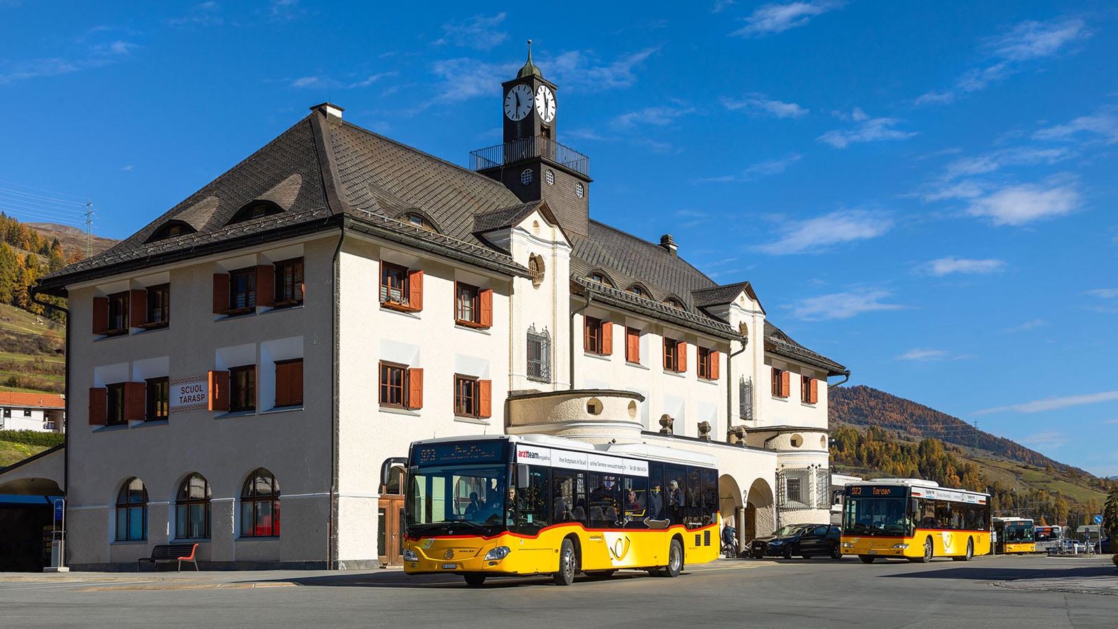 There are several yellow PostBus buses in front of the Scuol-Tarasp railway station building. The historic building with its clock tower and reddish-brown shutters is located in the sunshine, surrounded by hills coloured in autumn under a blue sky.