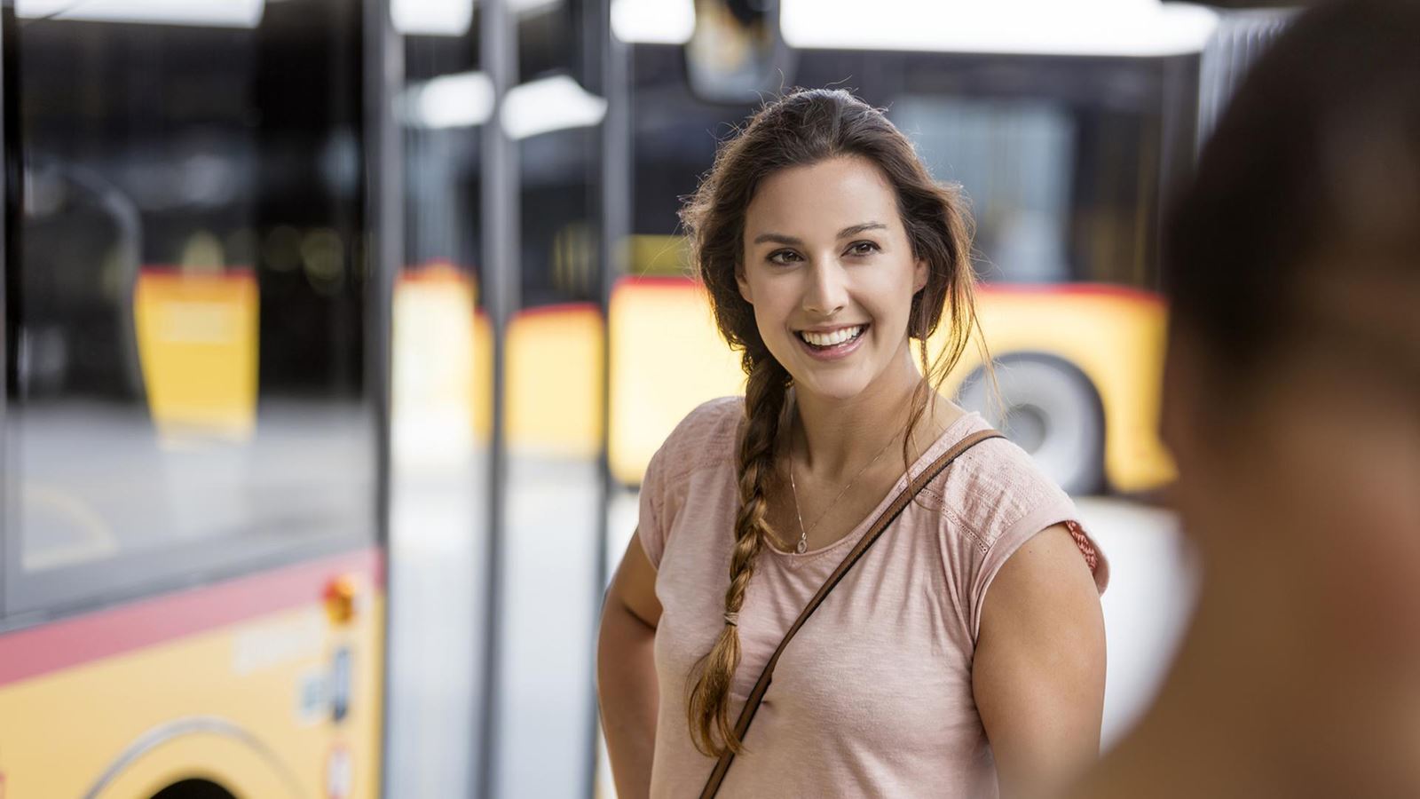 A smiling woman stands in front of a Postbus that is blurred in the background.