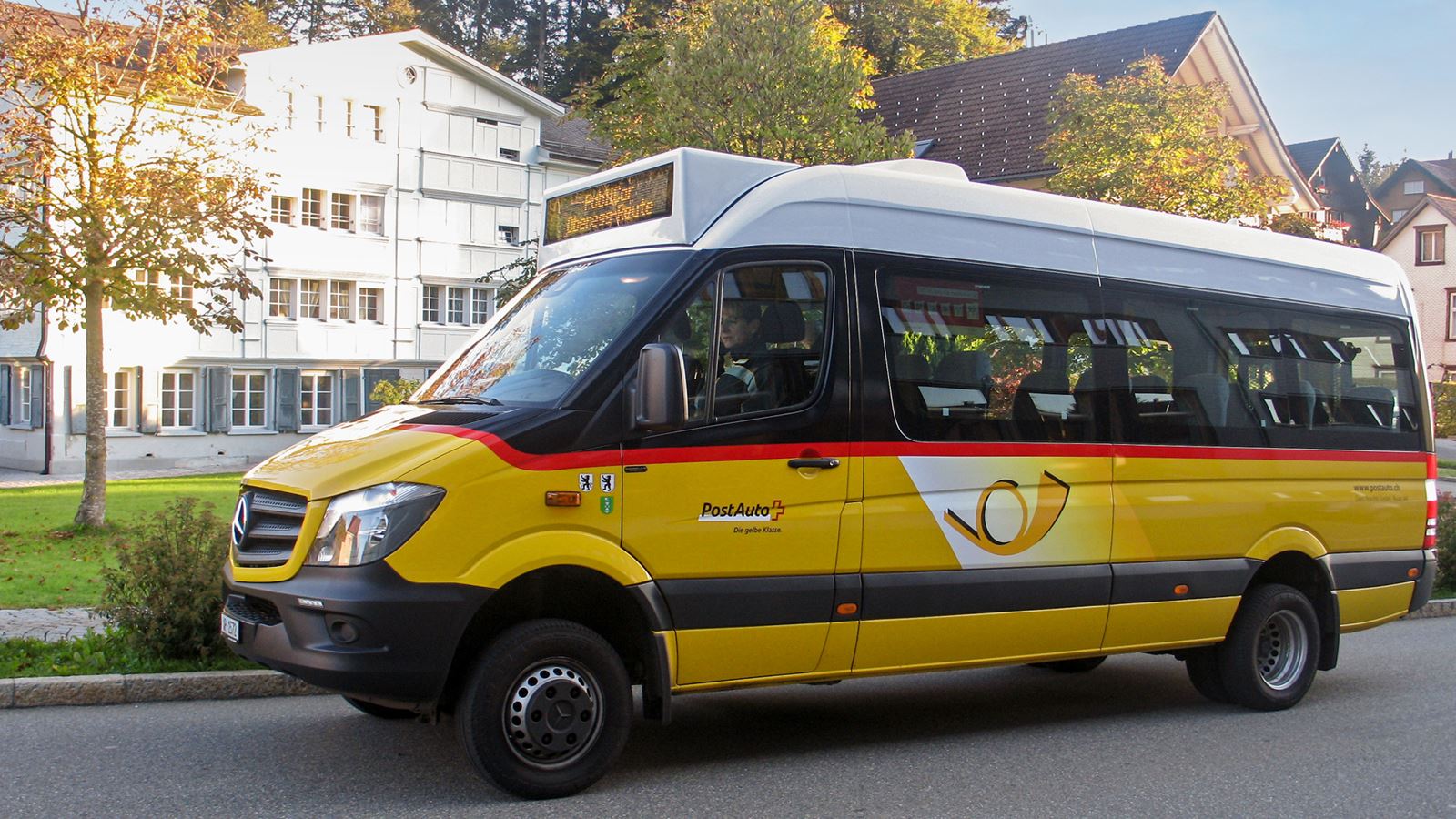 The small PostBus PubliCar shuttle on a hill next to a red bench. The Sun is setting through the mist in the background.
