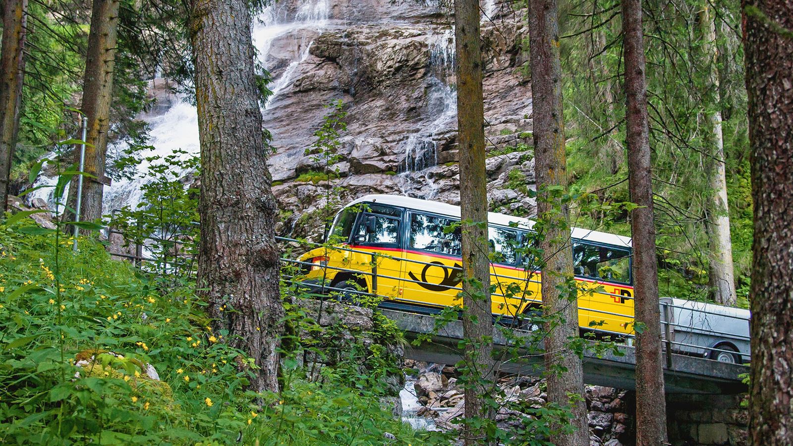 Kleines Postauto mit Anhänger fährt auf schmaler, steiler Strasse durch den Wald an einem Wasserfall vorbei.@Mike Niederhauser Merlin Photography 