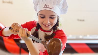 Una bambina con un cappello da chef firmato Maestrani versa col mestolo del cioccolato liquido in una pentola. © Maestrani Cioccolati Svizzeri SA