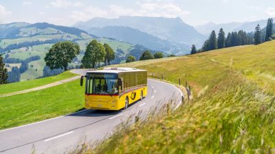 PostBus drives on roads in a rural area. In the background, the hilly landscape of Entlebuch with a view of the Schrattenfluh.
