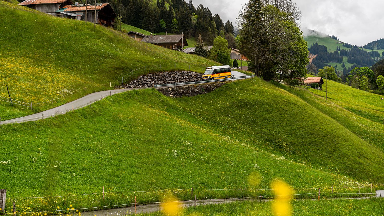 Kleines Postauto fährt die enge Strasse hinunter. Im Hintergrund sind traditionelle Häuser zu sehen und grüne Wiesen.