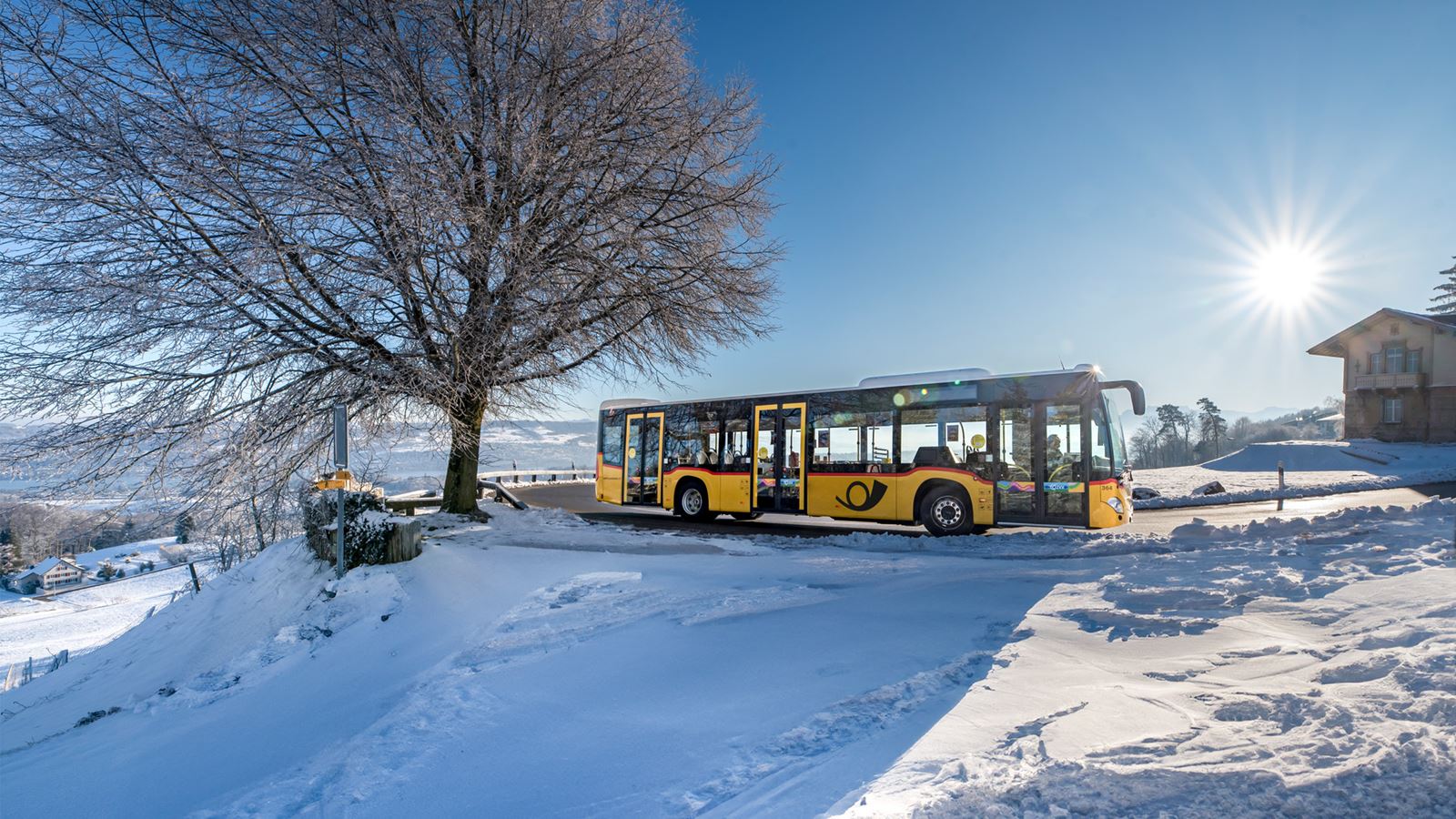 Ein gelbes Postauto fährt bei Sonnenschein durch die verschneite Landschaft auf dem Albispass.