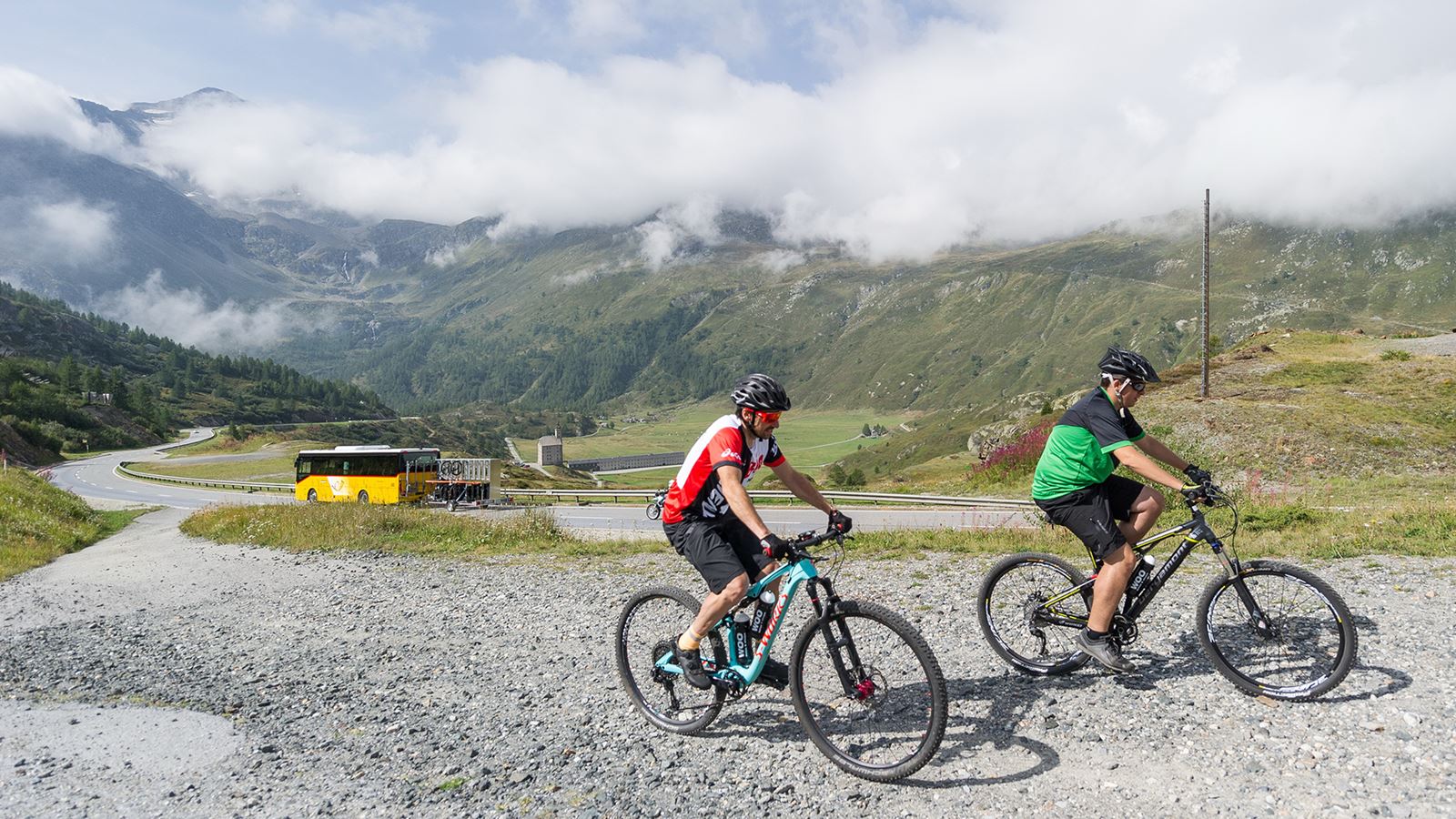 Two mountain bikers on the Simplon Pass. There is a Postbus in the background.