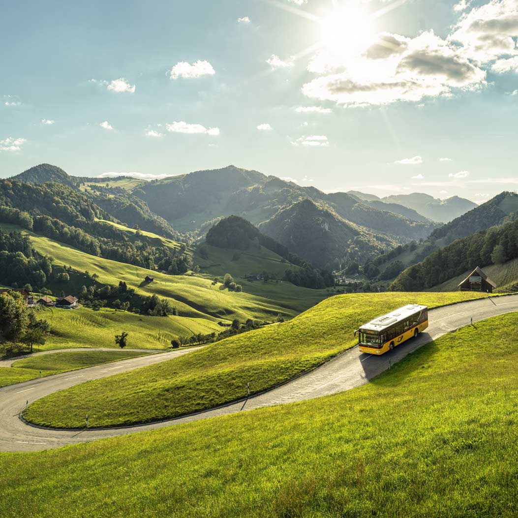 The Postbus drives down the passwang. The green, hilly landscape can be seen in the background.