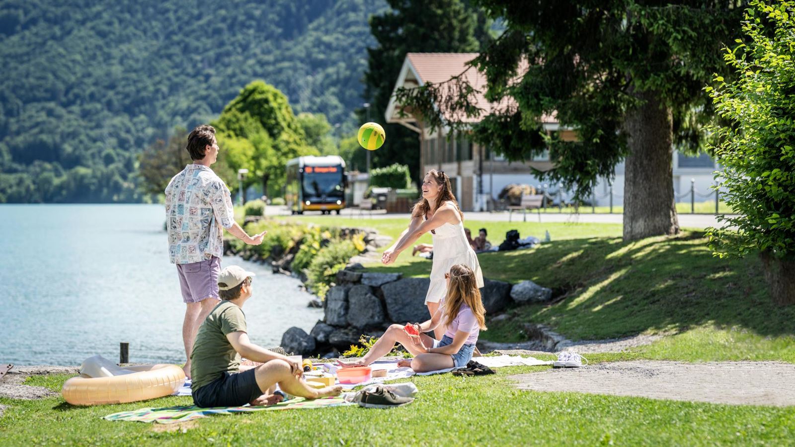Vier Personen machen ein Picknick am Seeufer und spielen mit einem Ball; im Hintergrund sind Bäume, ein Haus und ein Postatuo.