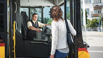 Eine Frau steigt auf der Fahrerseite in das Postauto ein. Der Chauffeur lächelt sie zur Begrüssung an.
