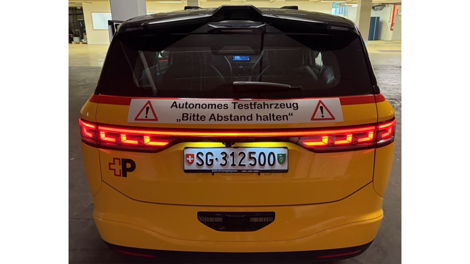 Ein gelbes AmiGo Fahrzeug von hinten. Über dem Nummernschild  ist ein Plakat mit der Aufschrift “Autonomes Testfahrzeug, Bitte Abstand halten!”  angebracht . 