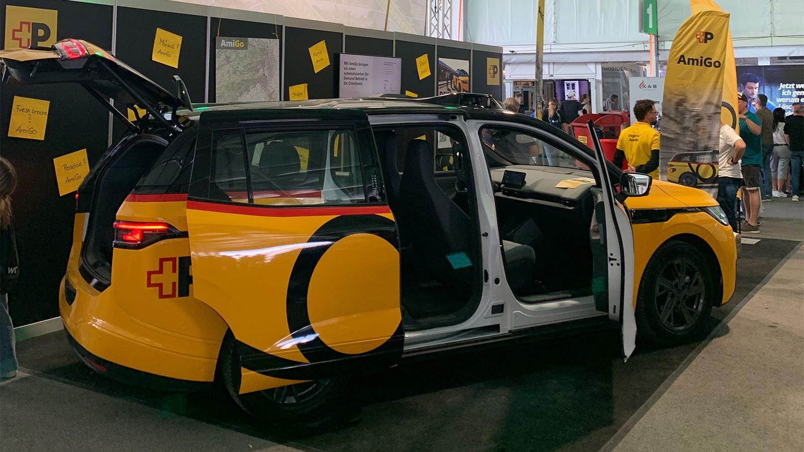 Gelbes AmiGo-Fahrzeug am Messestand steht mit geöffneten Türen in einer Halle; im Hintergrund Informationswände und Besuchende.