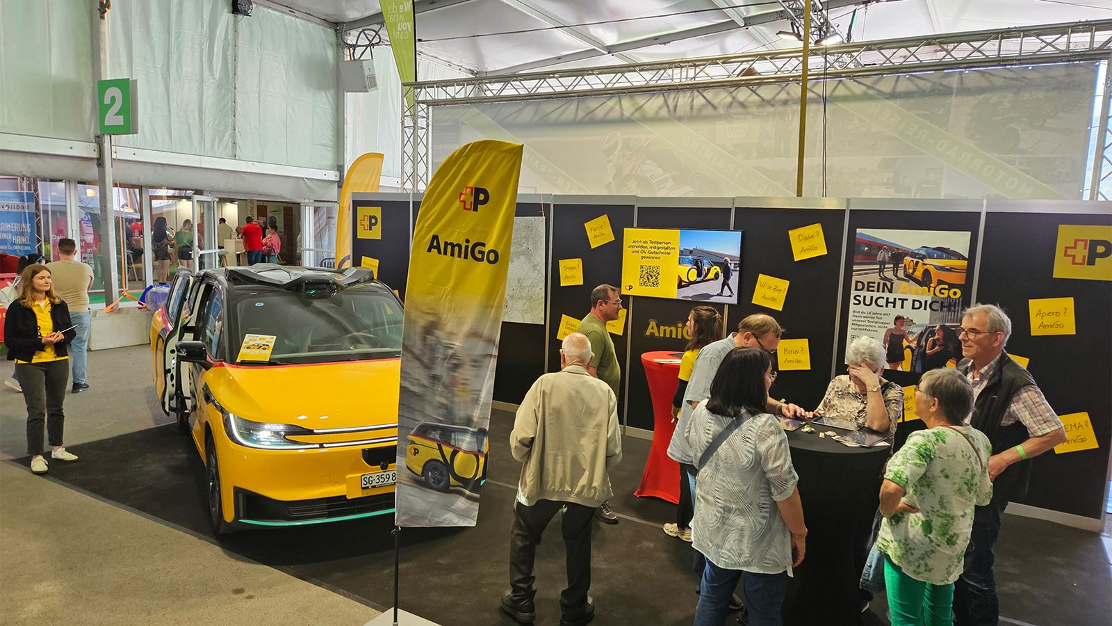 Le stand «AmiGo» avec un véhicule AmiGo jaune: plusieurs personnes s’informent sur des panneaux et des écrans.