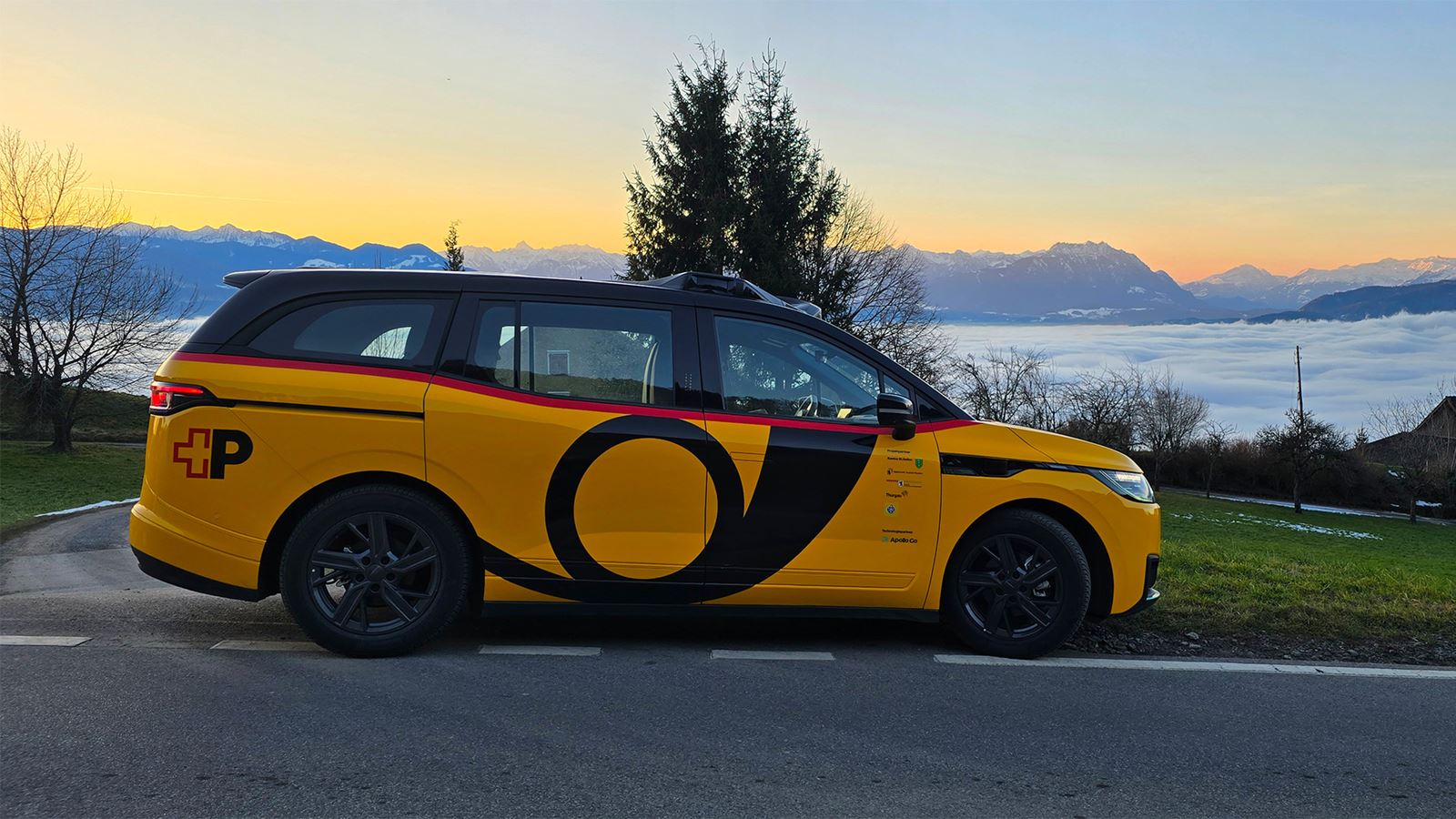 A yellow, autonomous AmiGo vehicle is standing at the roadside against a Swiss Alpine panorama with a sea of fog in the background.