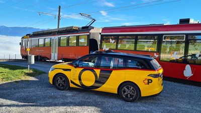 AmiGo Fahrzeug steht neben einem Zug an einer Bahnstrecke im ländlichen Raum.