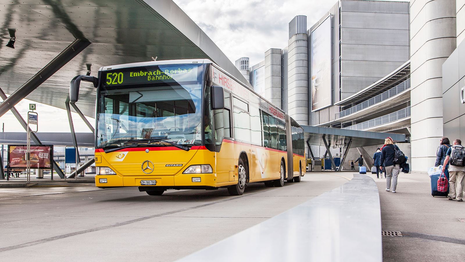 A Postbus stops at a train station in an urban setting.