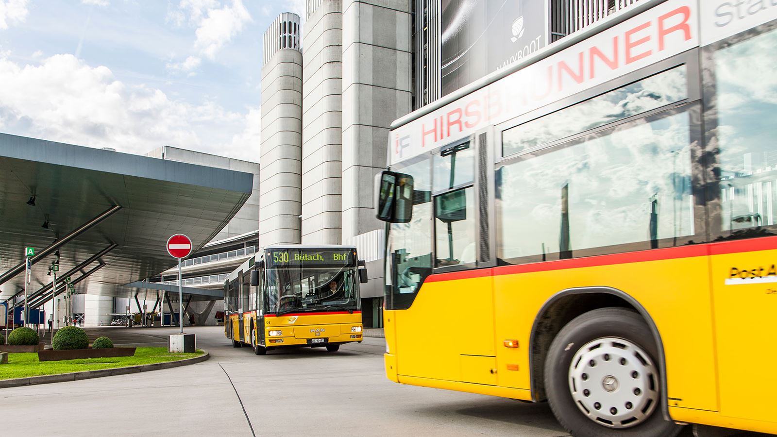 Two Postbuses intersect at a stop in front of a skyscraper.