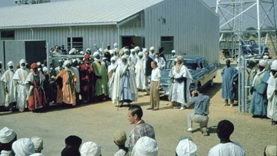 L’Emir de Kano et sa Cadillac entourés de nombreuses personnes portant des foulards et de longs vêtements.