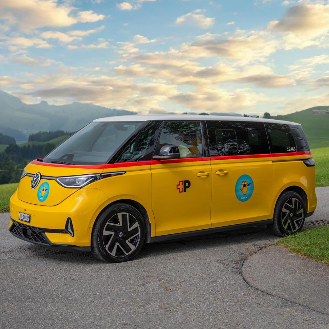 Elektrischer VW-Bus im gelben PostAuto-Design mit rotem Streifen und Posthorn-Logo, eingesetzt für den On-Demand-Service Publicar in Appenzell. Das Fahrzeug steht auf einer Landstraße in malerischer Schweizer Hügellandschaft, neben einem Wegweiser für Wander- und Fahrradwege
