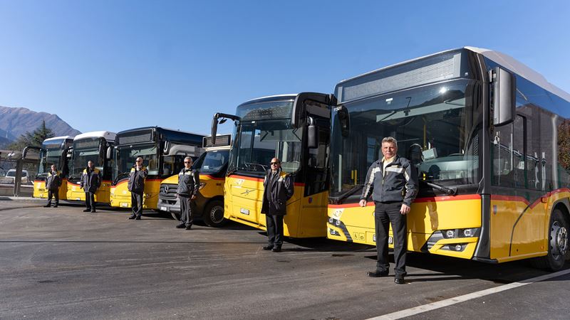 6 Postauto-Busse welche nebeneinander aufgereiht sind. Vor jedem Postauto steht ein Fahrer in Postauto-Uniform. 