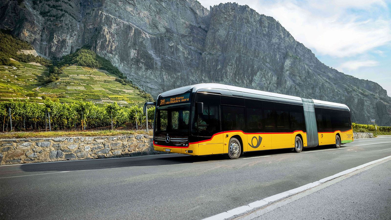 Ein elektrisches Gelenkpostauto auf einer Strasse, vor einem Berg, am Fusse von Weinbergen